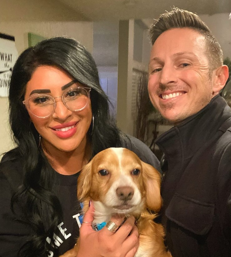 a smiling loving couple is holding a brown dog in their arms