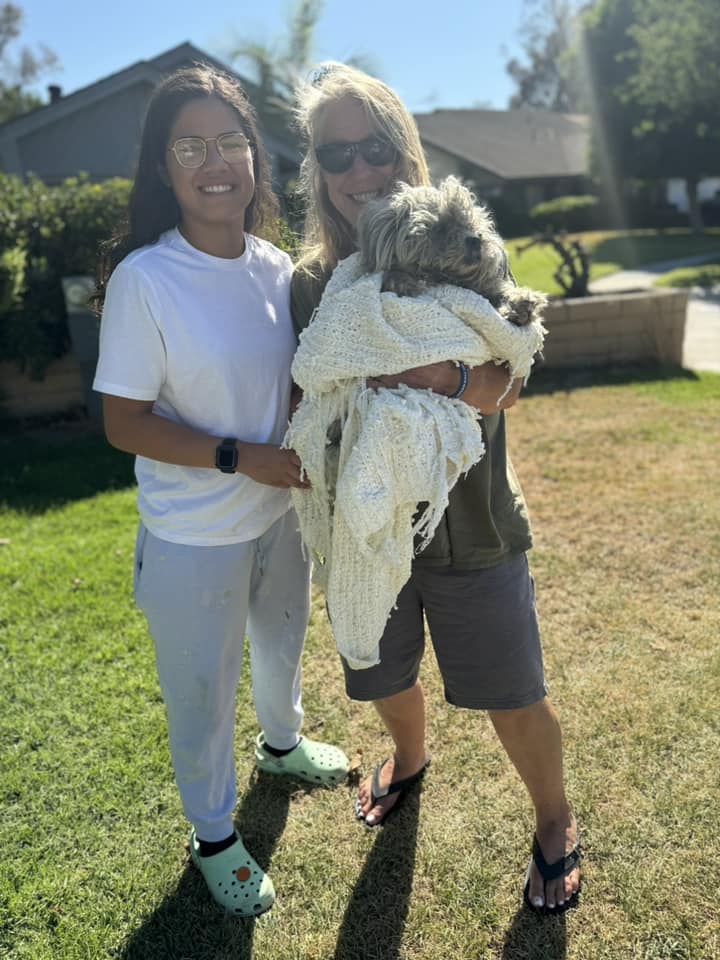 two smiling women are holding a dog wrapped in a blanket